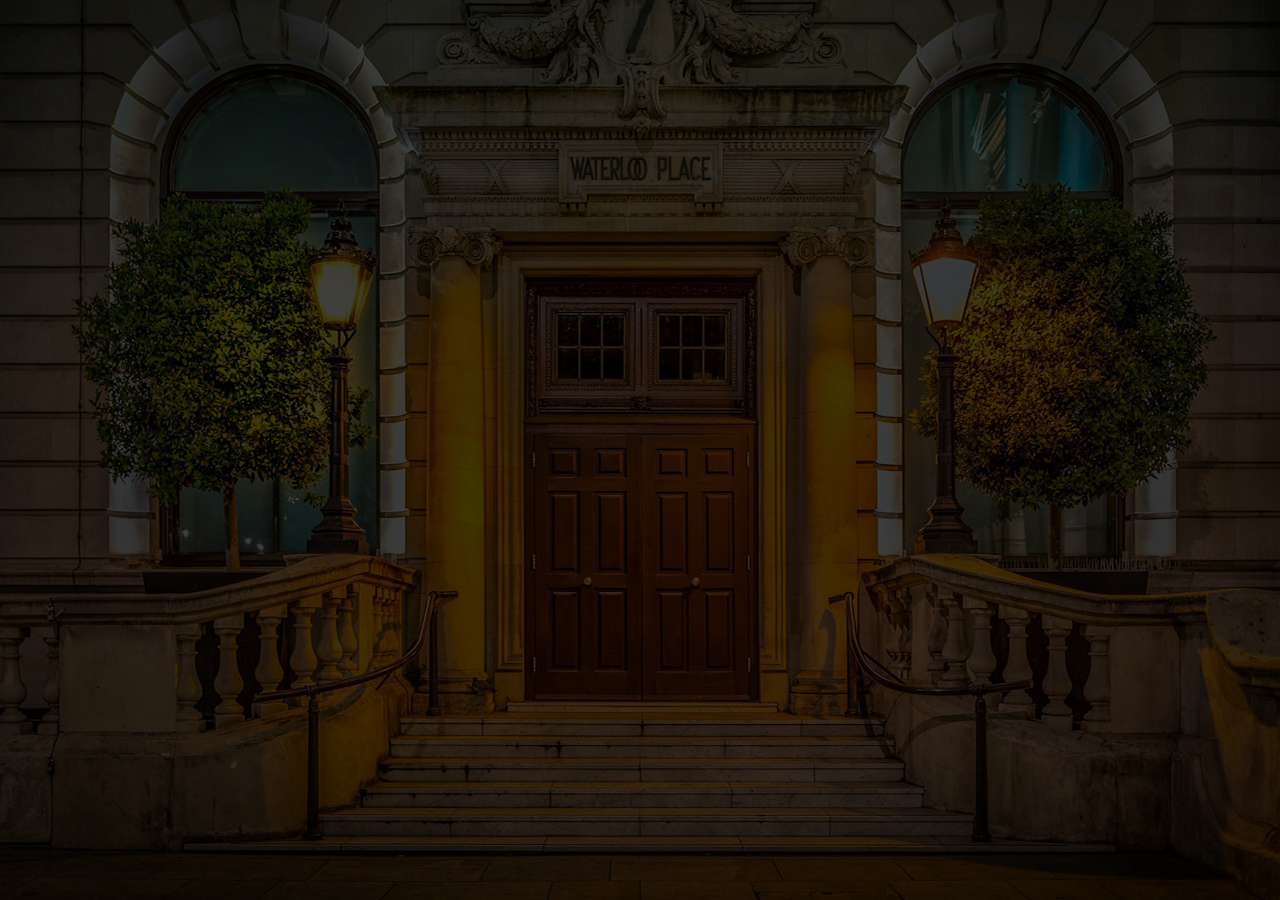 The entrance to a heritage building on Waterloo Place, Edinburgh with traditional heritage-style hardwood doors, with bushes and dimly lit street lanterns on both sides.