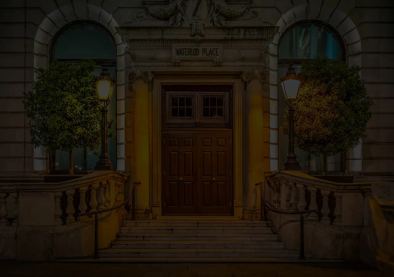 The entrance to a heritage building on Waterloo Place, Edinburgh with traditional heritage-style hardwood doors, with bushes and dimly lit street lanterns on both sides.