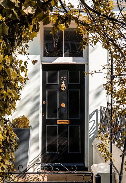 A Georgian-style timber black fanlight door, in a nature-focussed front yard, with polished brass fittings and the number 70.