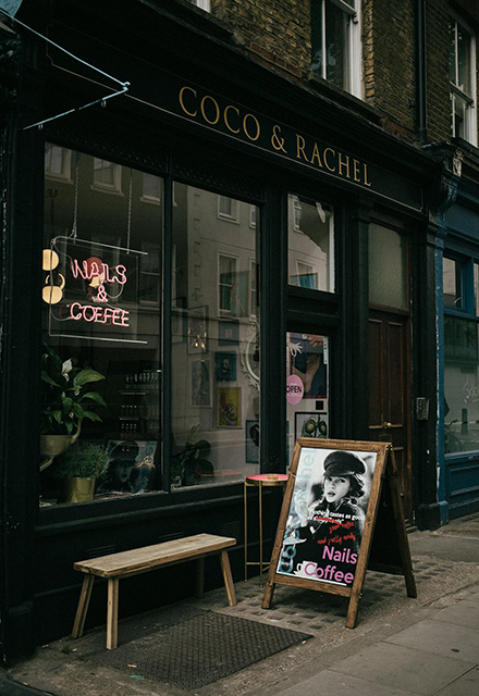A black shopfront on a high street with a small neon sign inside and an A-board outside.